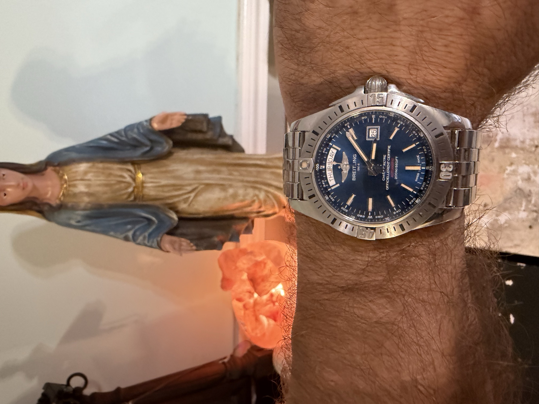 A steel Breitling Galactic 44 with a blue dial and day-date window, worn on
              David's wrist, with a statue of the Virgin Mary and a salt lamp behind it.