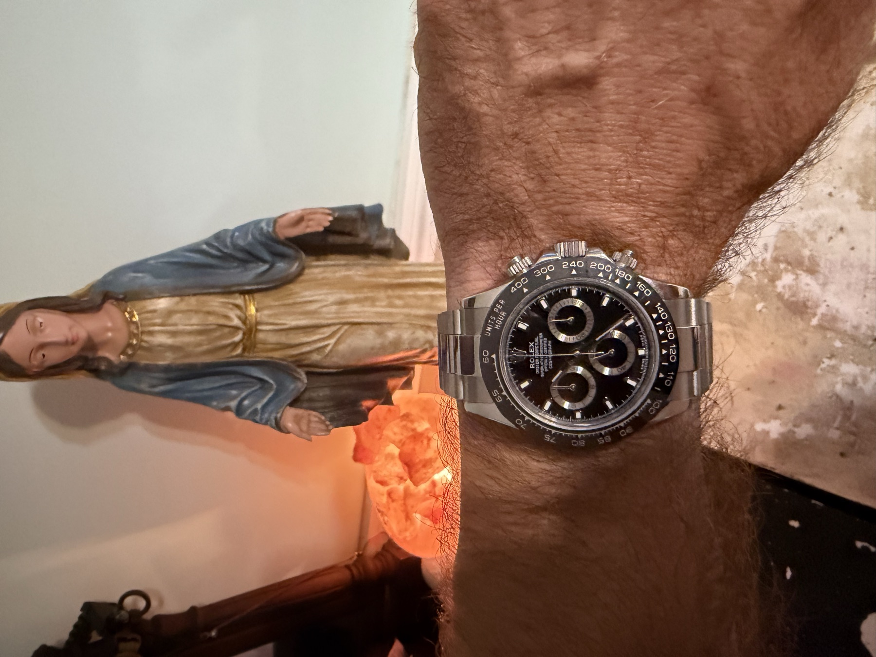 A steel Rolex Cosmograph Daytona on David's wrist, with a statue of the Virgin
              Mary and a salt lamp glowing behind it.